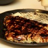 Para a finalização, coloque bastante molho para okonomiyaki, maionese, salpique aonori, katsuobushi e decore com benishoga